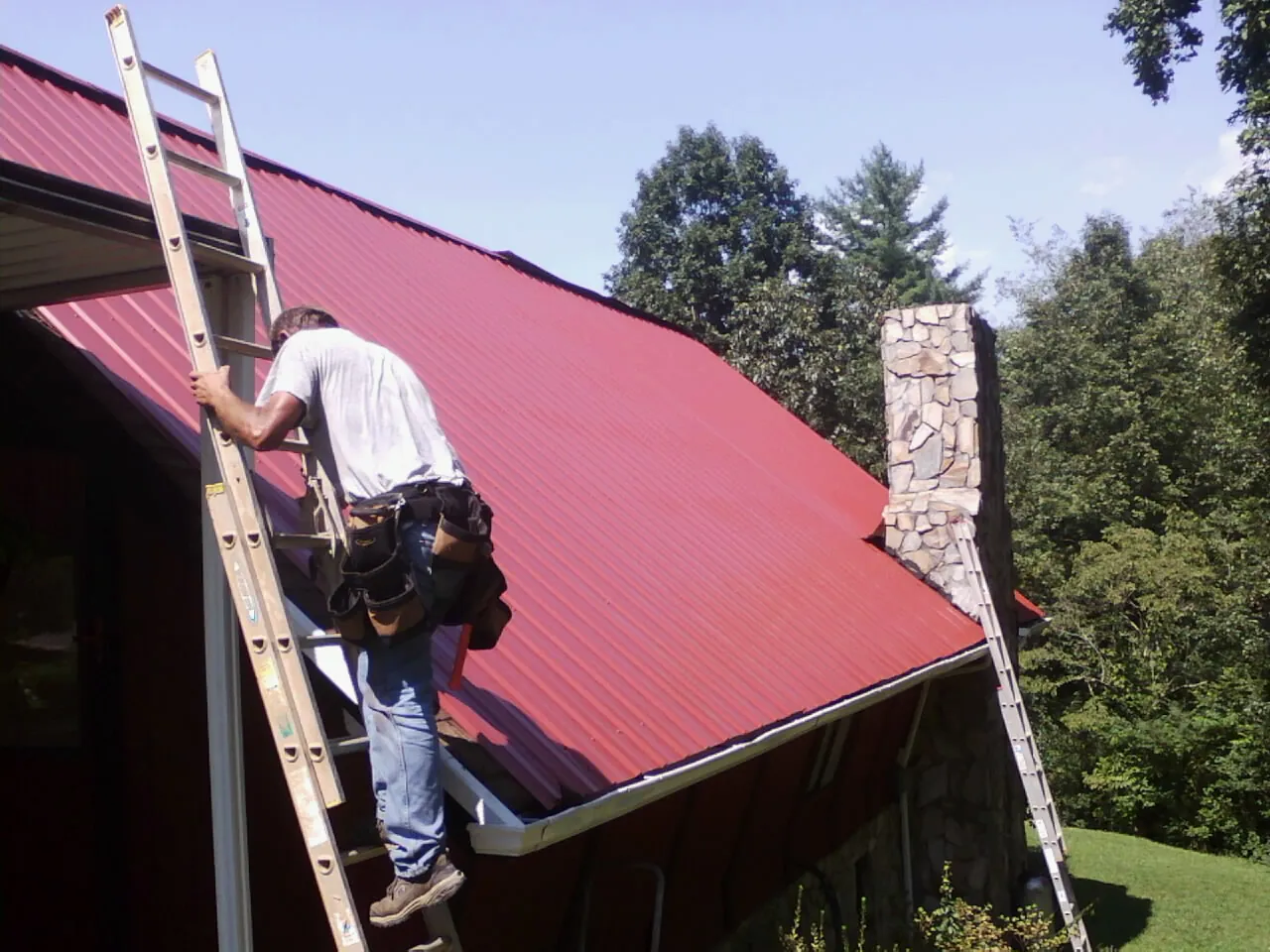 Roofer on a ladder beside a red metal roof and stone chimney — Soffit & Fascia Repair by Roofing Brothers in Loma Mar