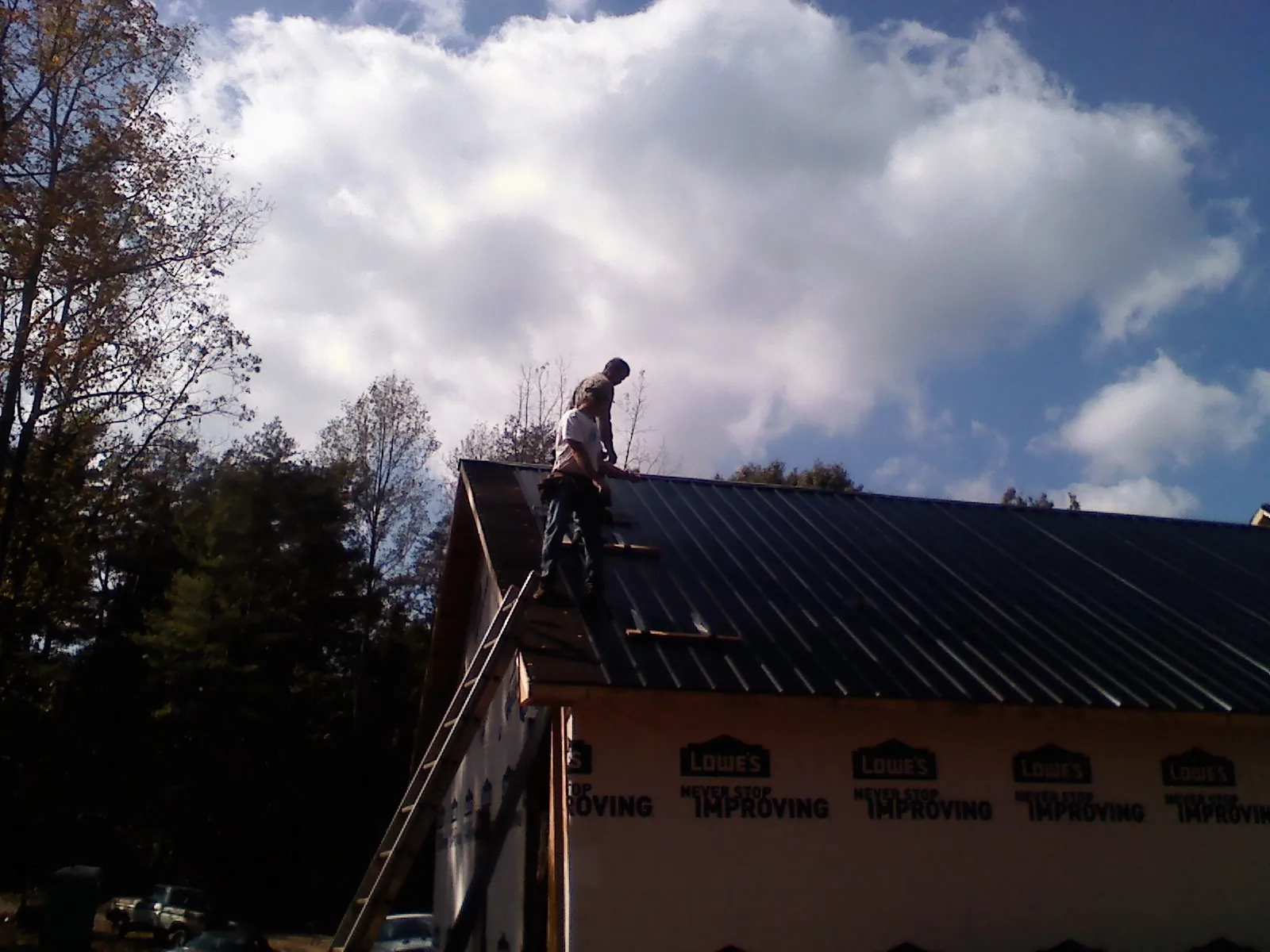 Roofer fastening dark metal panels high against a cloudy sky, from a Roof Inspection job in Loma Mar